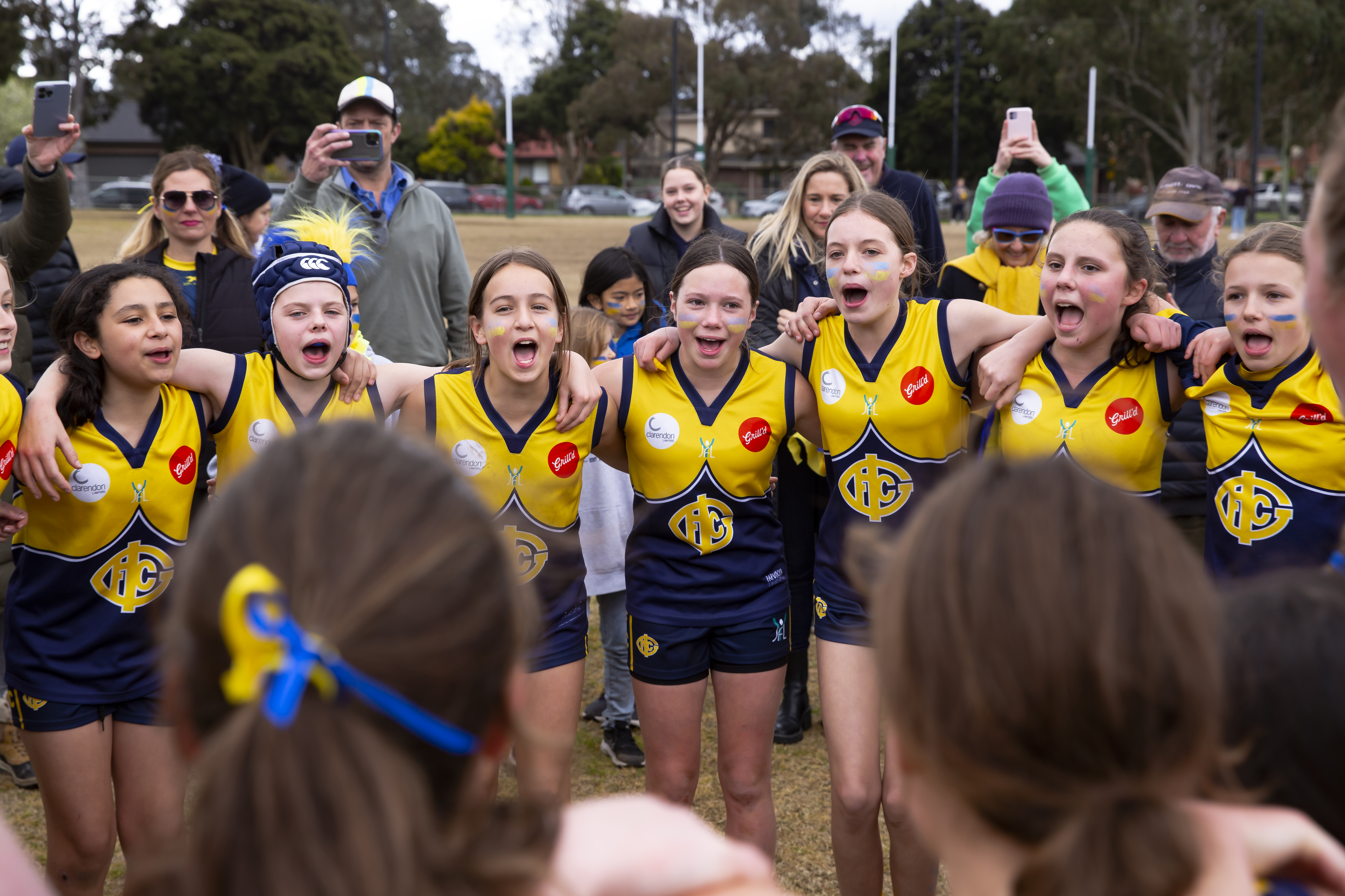 Young girls from the Yarra Junior Football League stand arm in arm celebrating a win. They are in the middle of singing their club song.
