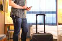 Man using smartphone in hotel room with baggage and suitcase. Tourist with mobile phone in holiday rental apartment. Calling taxi cab or doing online check-in.