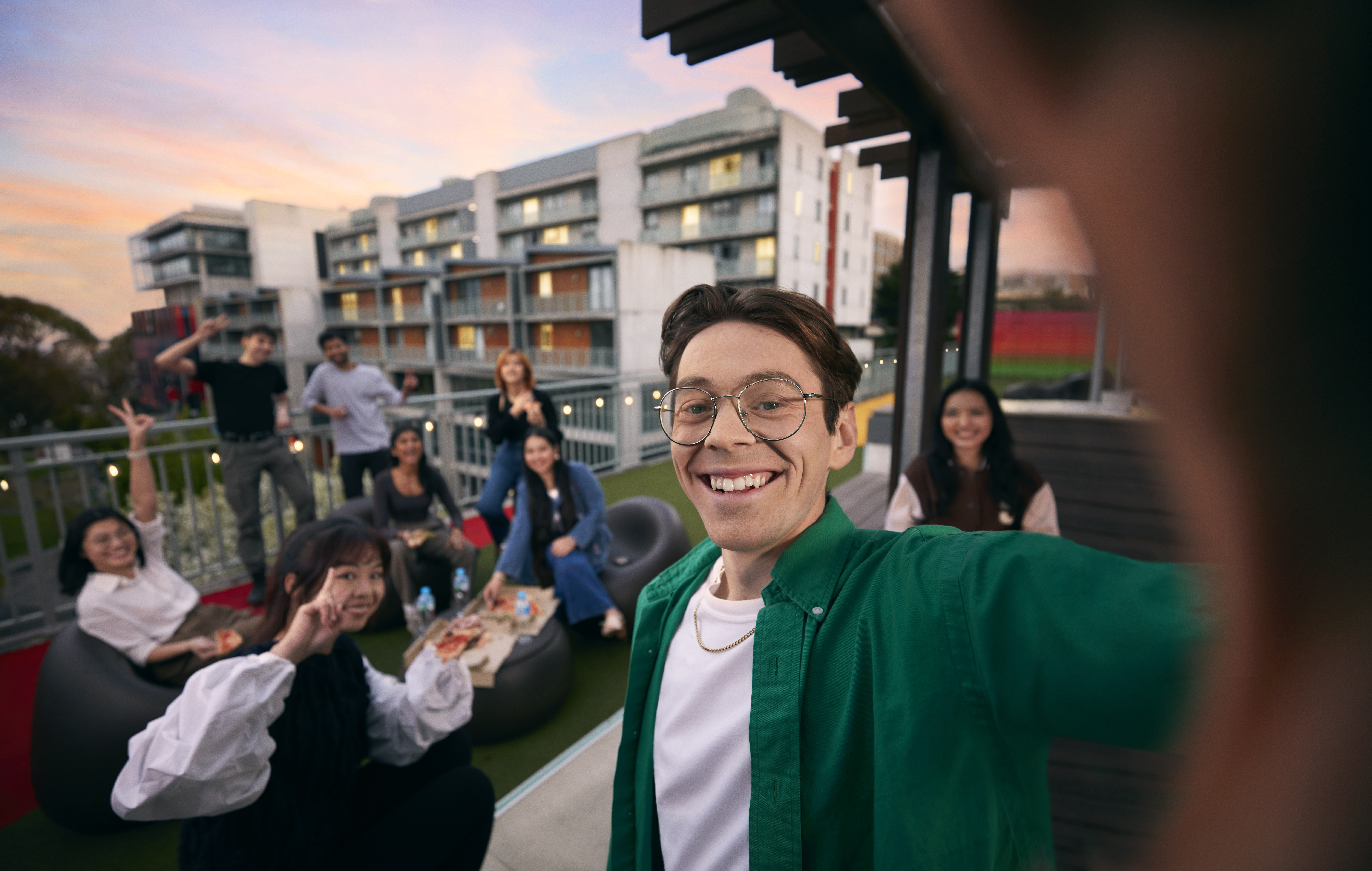Male student ambassador is taking a photo in selfie style, with group of friends smiling in the background. They are on a balcony eating food with the Hawthorn campus in the background. 