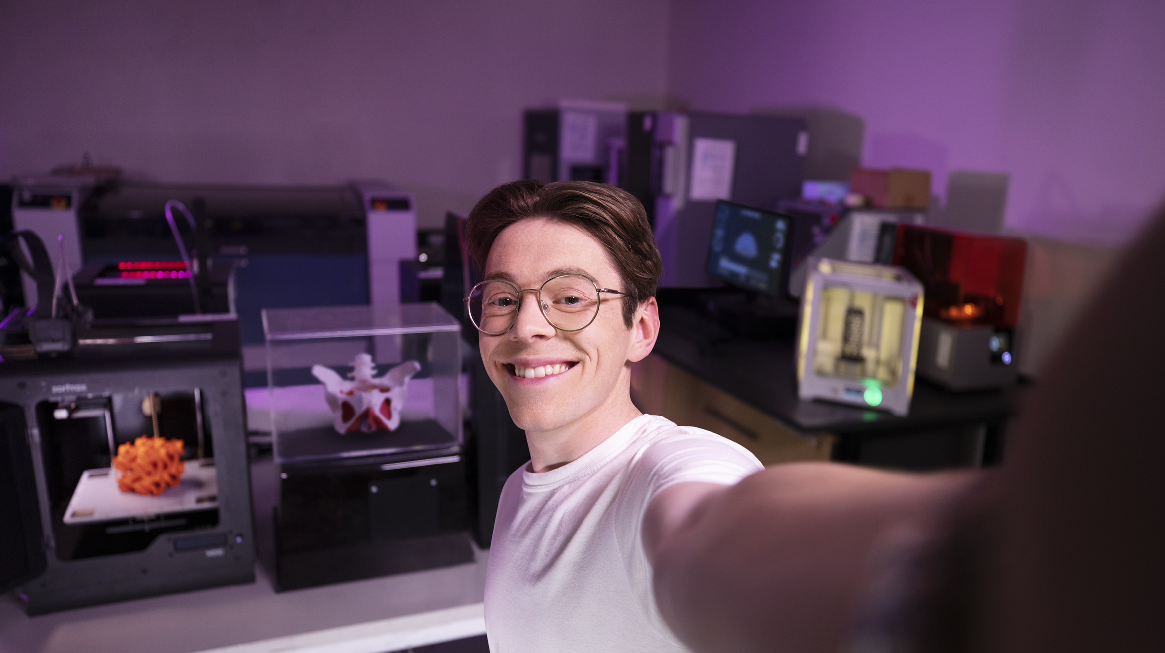 Male student smiling at the camera taking a selfie style photo, in the background shows a dimly lit science lab. 