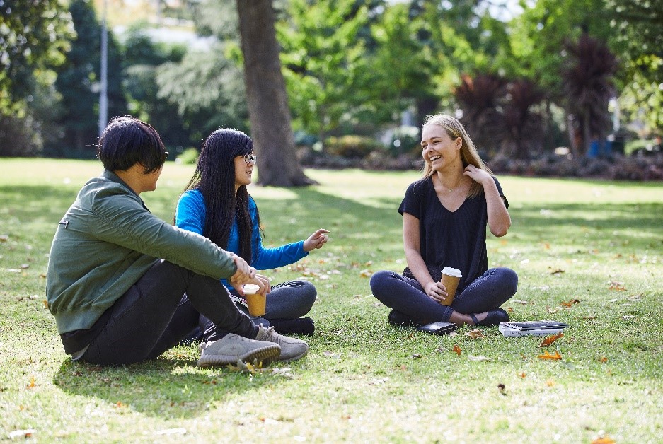 Three students sit in the park laughing and drinking coffee