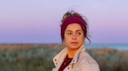 Close up of a young woman wearing a maroon head band and tan jacket, staring wistfully off into the distance.