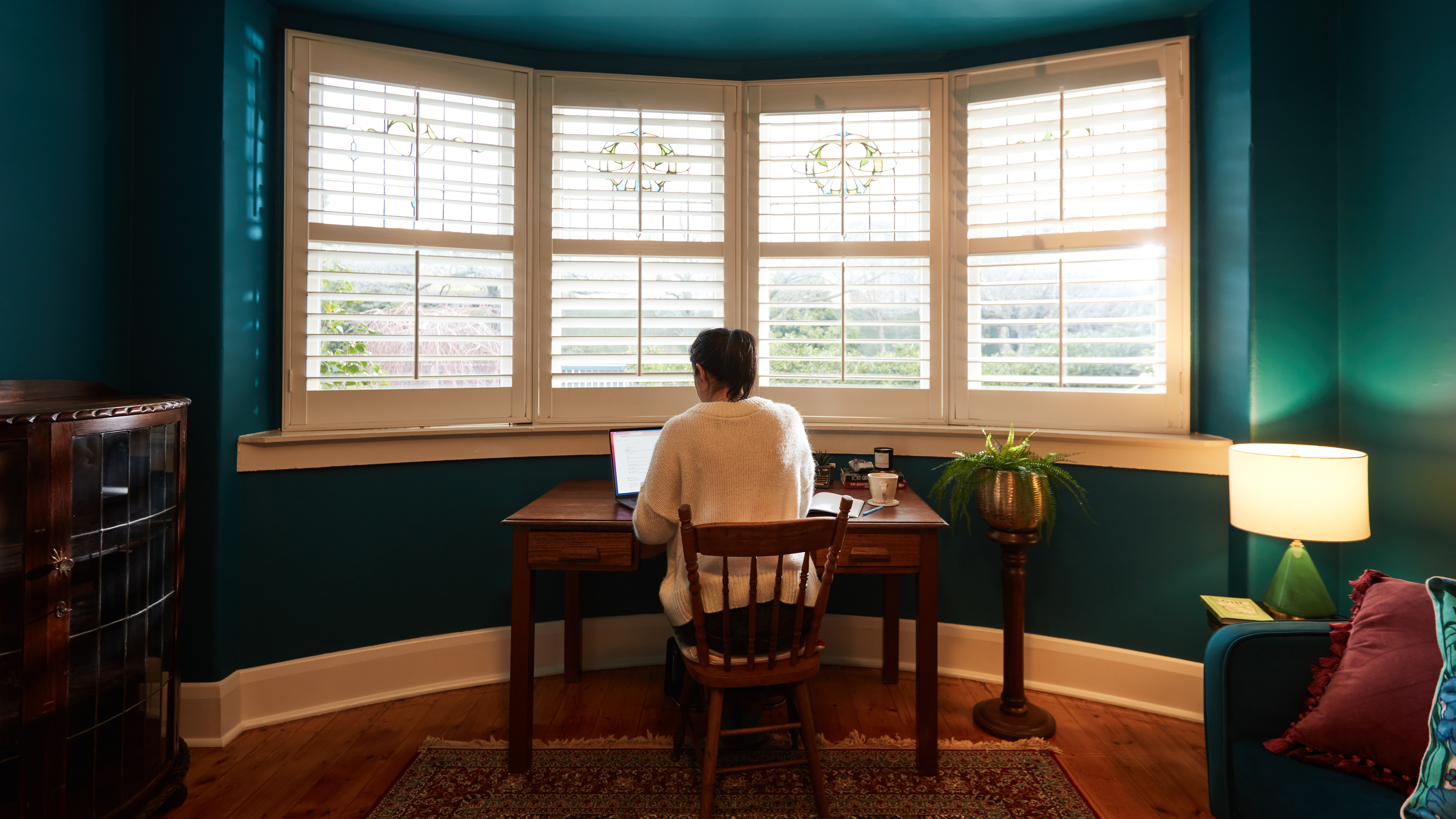 A student studying at an old wooden desk by a bay window. Daylight streams through and casts onto turquoise-painted walls..