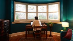 A student studying at an old wooden desk by a bay window. Daylight streams through and casts onto turquoise-painted walls..