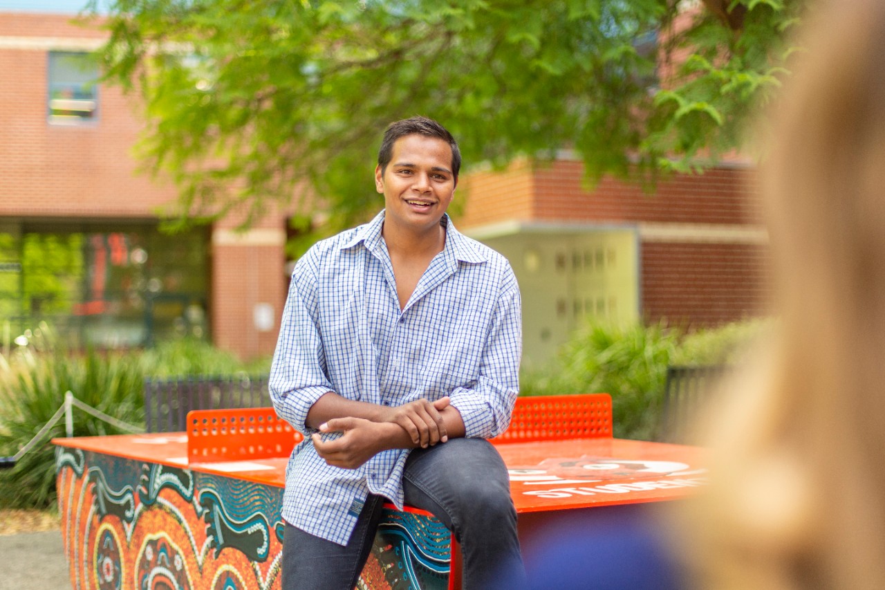 An Indigenous student sitting on an outdoor table tennis table decorated in Swinburne's Reconciliation Artwork on the Hawthorn campus