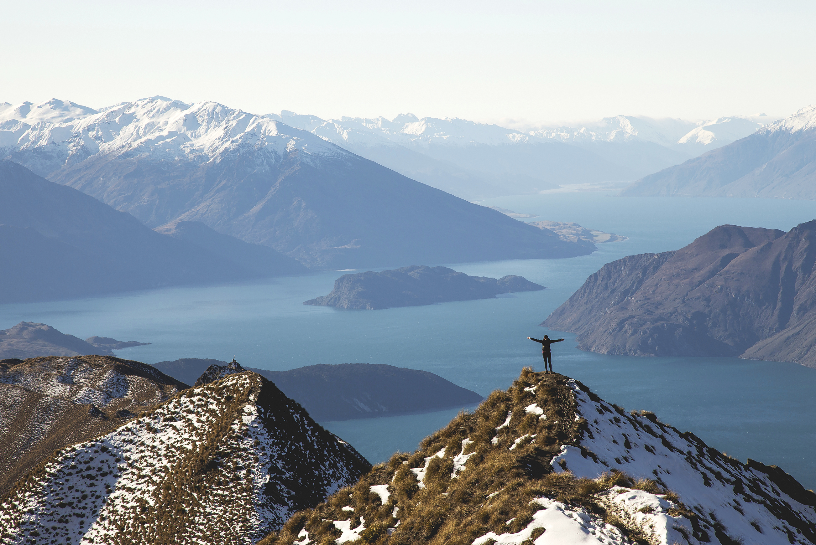Person stands on a summit peak, arms outstretched, looking out to mountains and the sea.