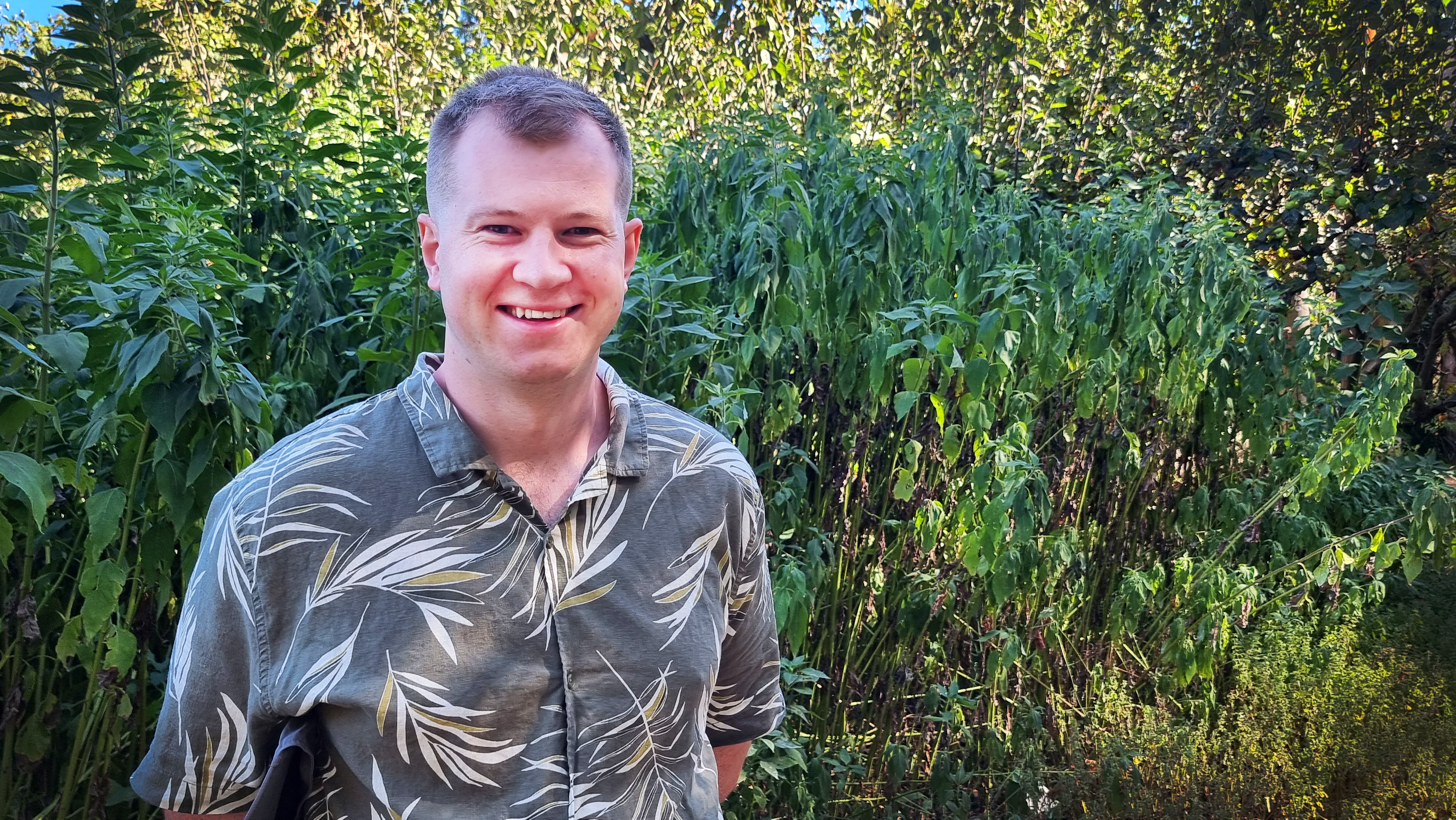 Leith wears a brown collared shirt with leaf patterns and stands in front of tall shrubs and trees 