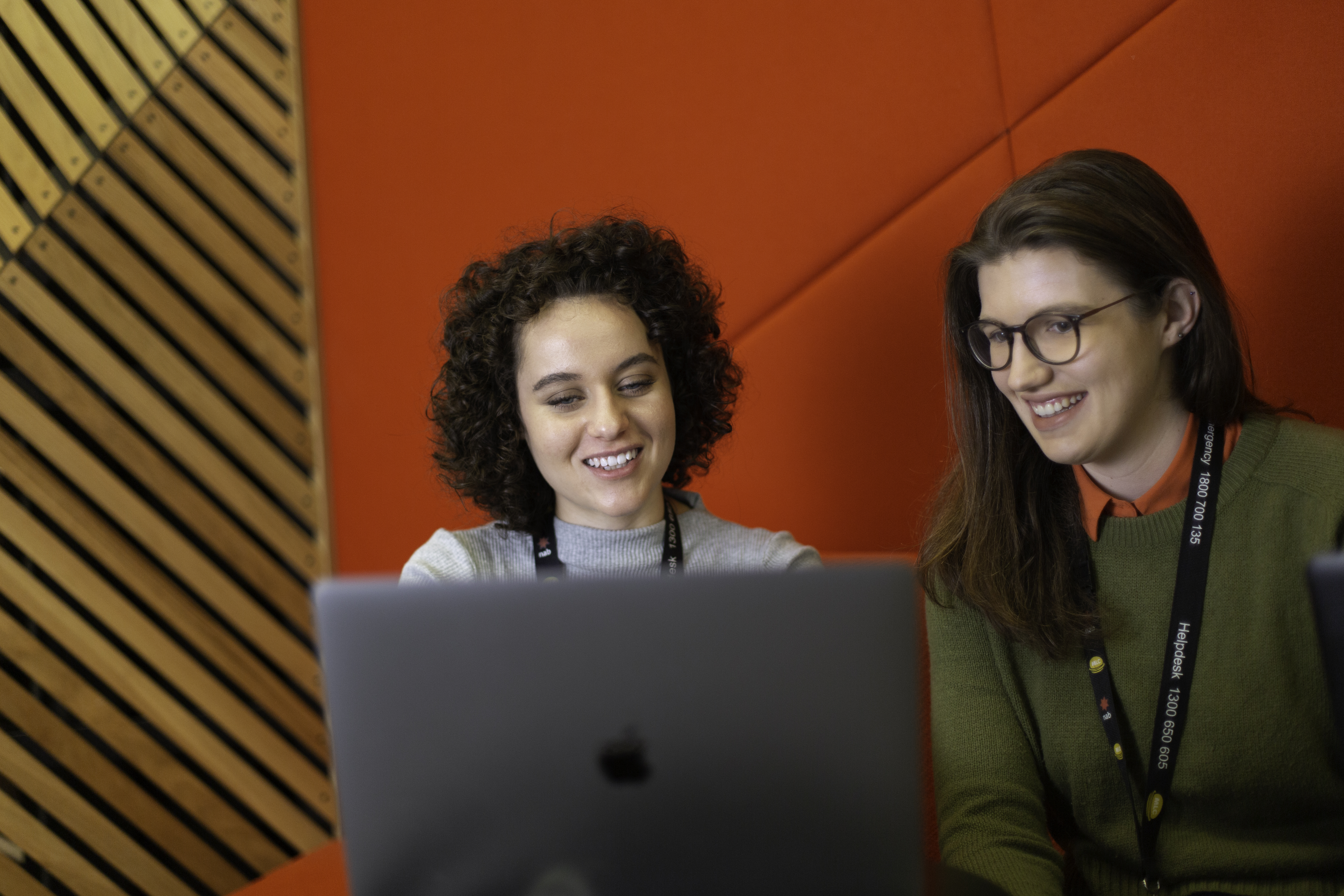 Two female students working at NAB sit on a red chair and view a presentation on a laptop