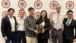 Five students and an academic stand in front of a photo wall holding a plaque  