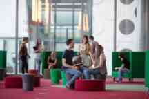 A group of undergraduate students talk inside a building on campus sitting on stools laughing smiling