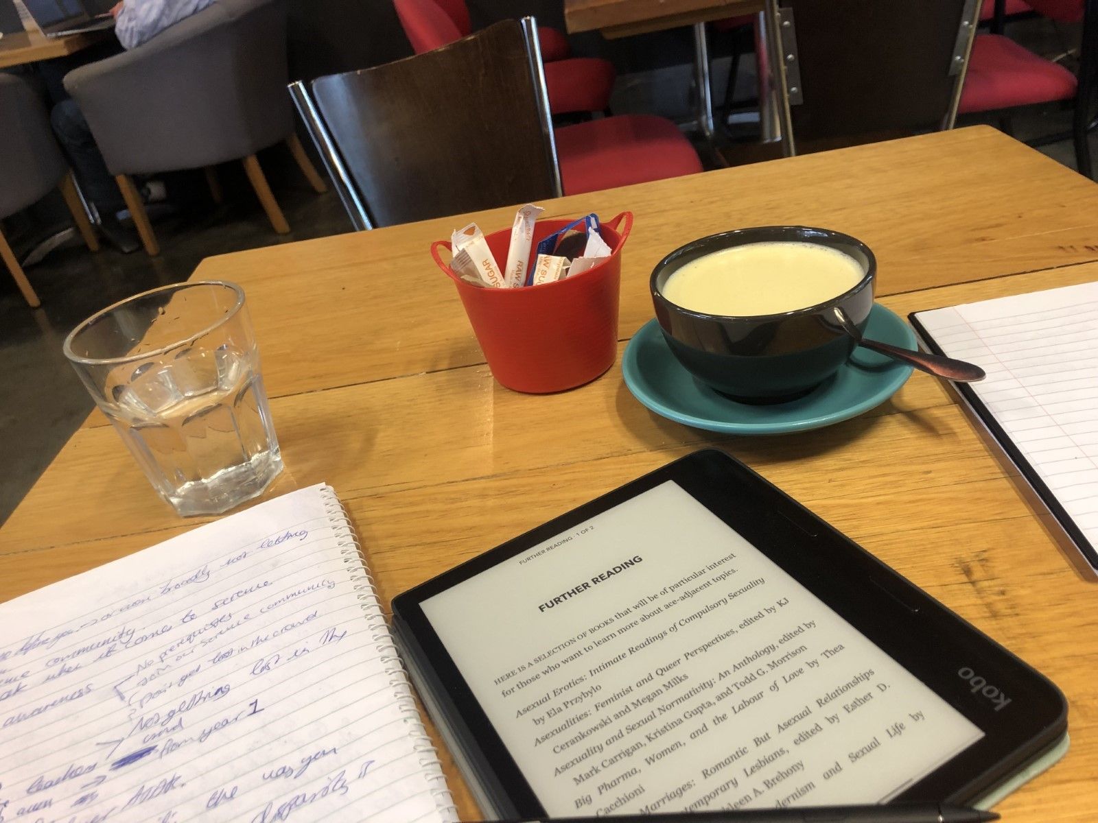 Notebooks and an e-reader strewn across a cafe table alongside a turmeric latte. 