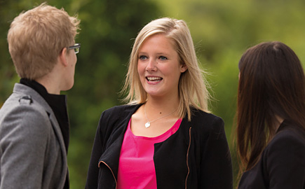 Three students are having a conversation outside. The camera focuses on a blonde lady wearing a black jacket and pink top.